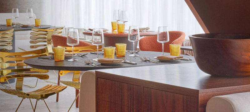 Ambiente interno do Baio Cozinha Sulista, com mesas postas, taças, louças e cadeiras em diferentes acabamentos. Grandes janelas com cortinas claras iluminam o salão, que apresenta decoração contemporânea em tons terrosos e detalhes dourados.