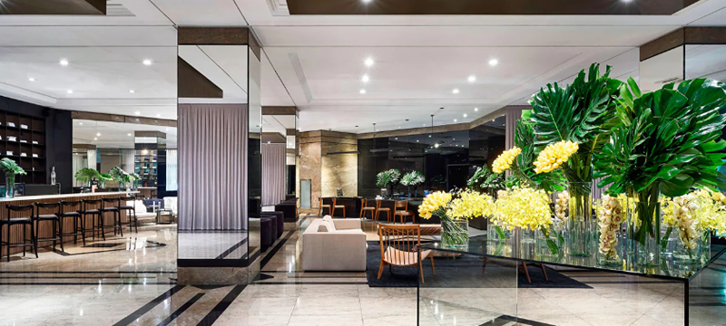 Lobby do Sheraton São Paulo WTC Hotel, com amplo salão, áreas de convivência e arranjos de flores e folhagens sobre uma mesa de vidro. O ambiente apresenta piso de mármore, iluminação embutida e mobiliário contemporâneo.