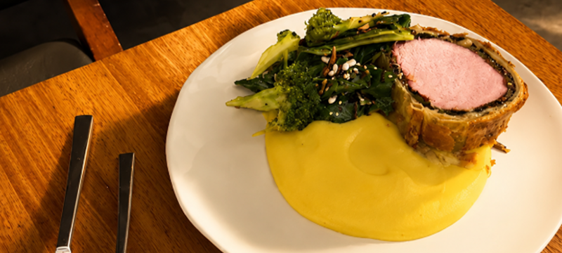 Prato de Beef Wellington servido com purê de mandioquinha e vegetais verdes em uma mesa de madeira do Terraço Notiê by Priceless.