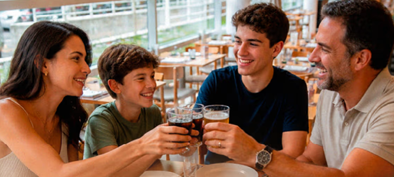 Família reunida à mesa no CipóRestô, com dois adultos e dois jovens compartilhando uma refeição em um ambiente iluminado por grandes janelas.