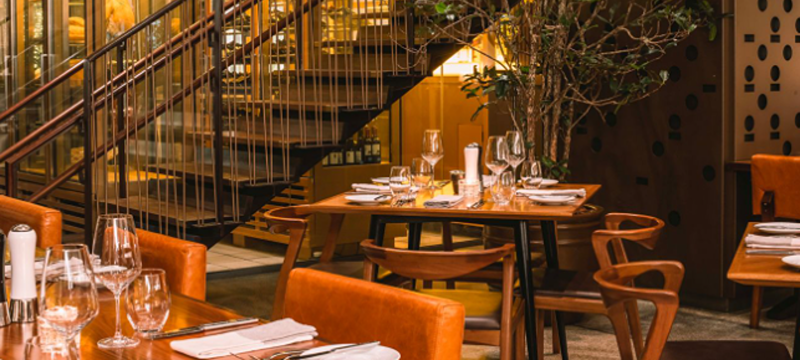 Ambiente interno do Restaurante Neto, com mesas de madeira, cadeiras em tons terrosos e escada ao fundo.