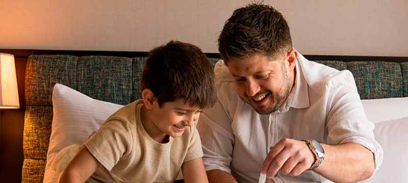 Pai e filho sentados sobre uma cama, sorrindo enquanto abrem juntos uma caixa de presente.
