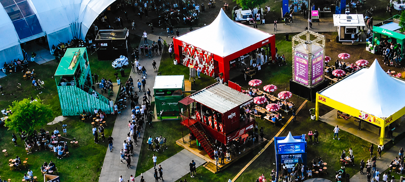 Vista aérea do festival SP Gastronomia ao ar livre, com circulação de público entre tendas, estandes e espaços gastronômicos. Mesas com guarda-sóis e diferentes estruturas.