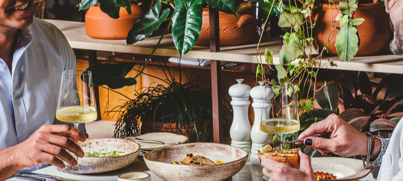 Duas pessoas compartilham uma refeição em uma mesa do Restaurante Neto. Pratos com diferentes preparações, pães e taças com bebidas compõem a mesa, cercada por plantas e vasos que integram a decoração do ambiente.