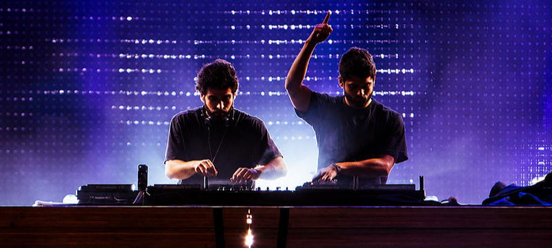 Dois DJs se apresentam em um palco, atrás de uma mesa de equipamentos. Um deles opera a mesa de som enquanto o outro levanta um dos braços. Luzes em tons de roxo e azul e uma forte iluminação ao fundo compõem o cenário.
